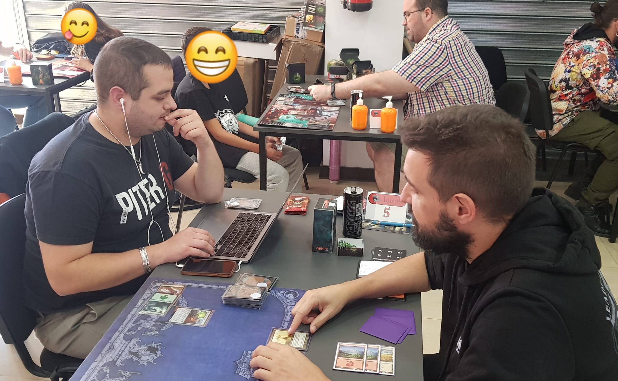 Magic Accesible VS Tasigur en la tienda de El Túmulo. Detrás aparecen Grego Jr, con la cara tapada por un emoji al ser menor de edad, jugando contra Tiamac. También aparece el ordenador de Magic Accesible y las cartas en la mesa